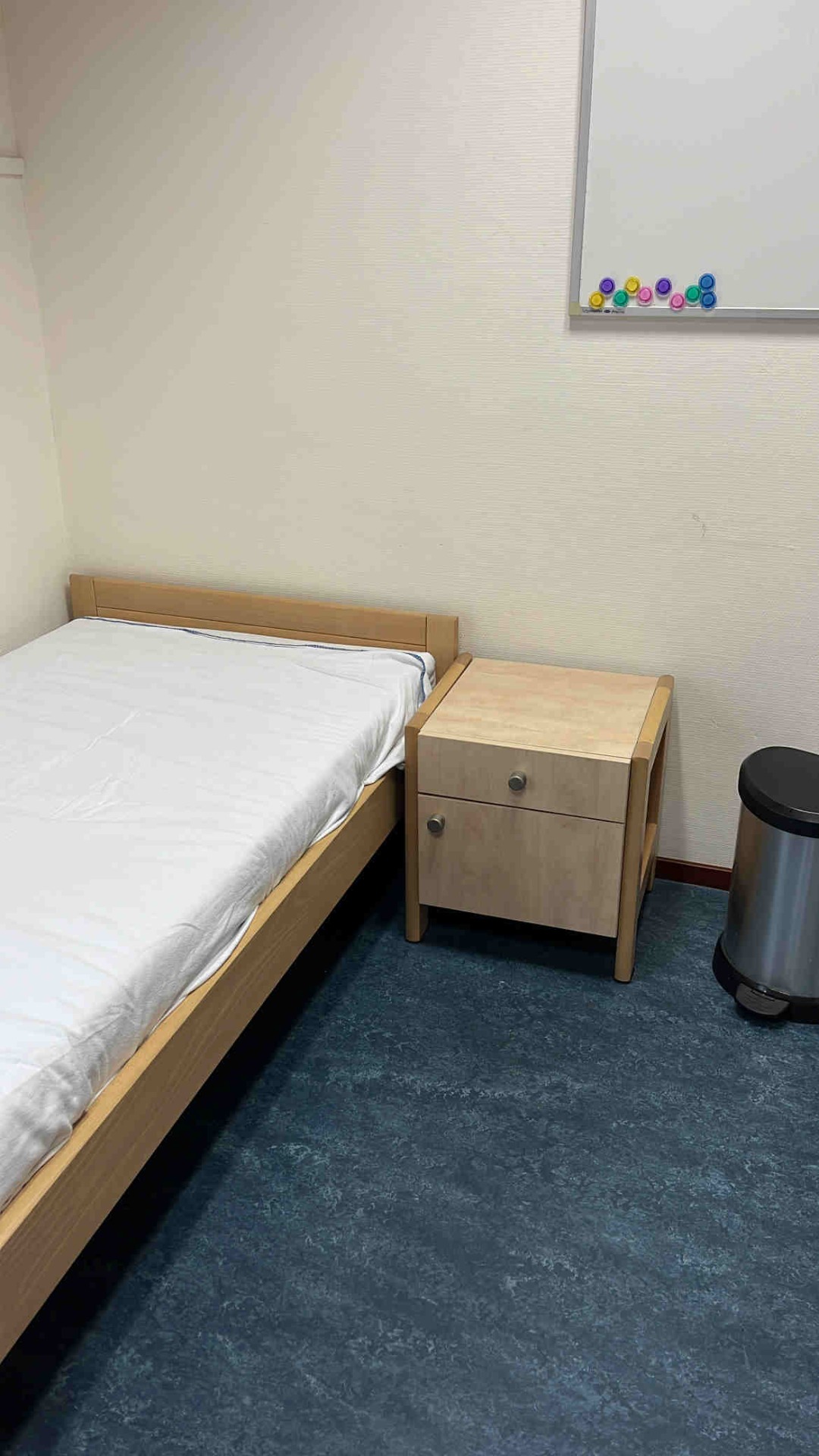 Een eenpersoonsbed met houten frame, nachtkastje met lade, en een whiteboard met gekleurde magneten. Crisisopname in een kliniek.