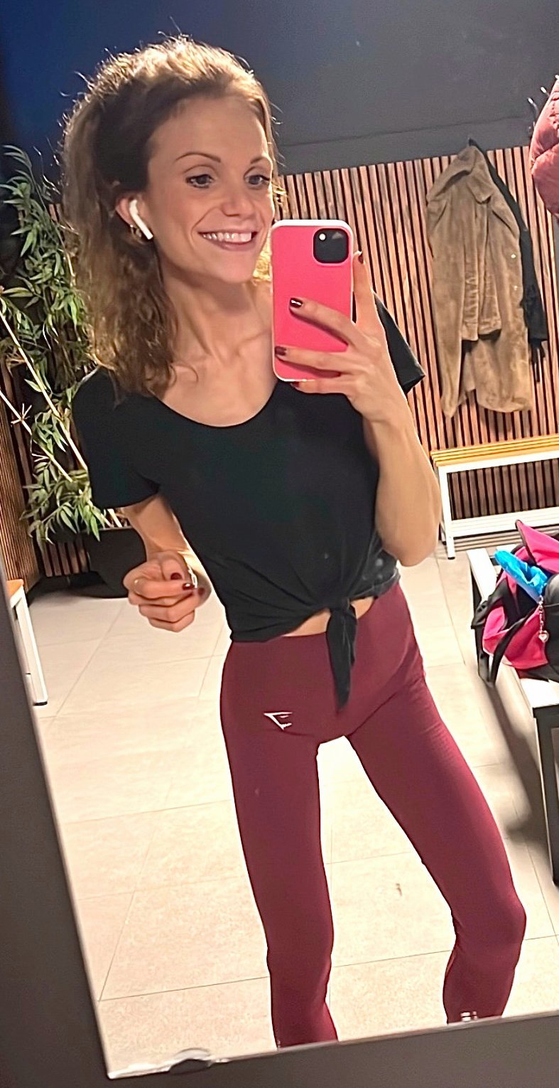 Vrouw maakt selfie in spiegel in de sportschool na een goede sportsessie, draagt zwarte top en bordeaux legging, buiten balkon met kledingrekken.