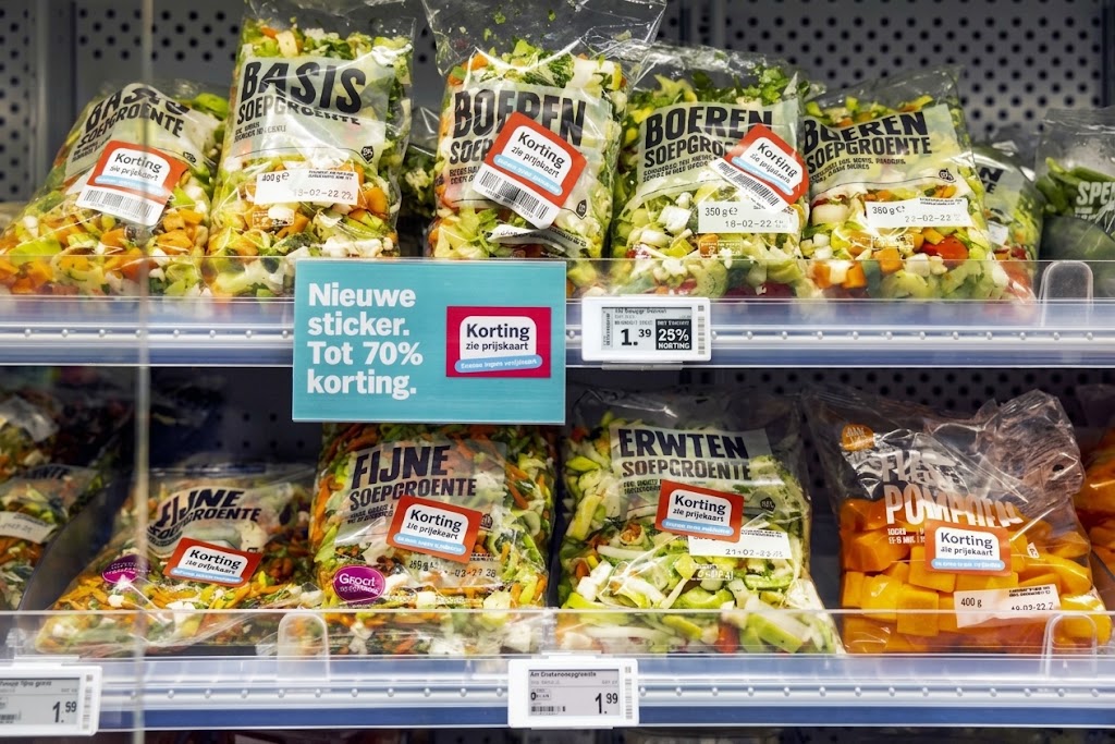 Rek met verpakte soepgroenten en kortingsstickers in een supermarkt, met prijskaartjes en promotiebord.