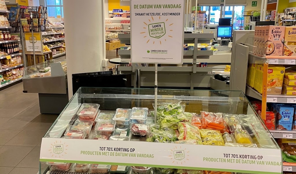 Supermarkt met kortingsdisplay en gekoelde maaltijden, borden met “De datum van vandaag” en “Samen minder verspillen”.