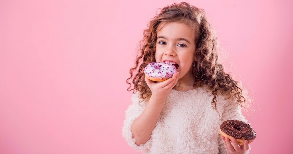Jong meisje met krullend haar houdt een donut vast en glimlacht, met roze achtergrond en tweede donut.
