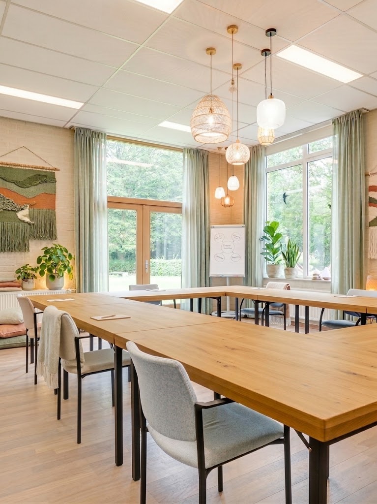 Ruime vergaderruimte of therapieruimte met houten lange tafel, grijze stoelen, hanglampen en grote ramen met groen uitzicht.