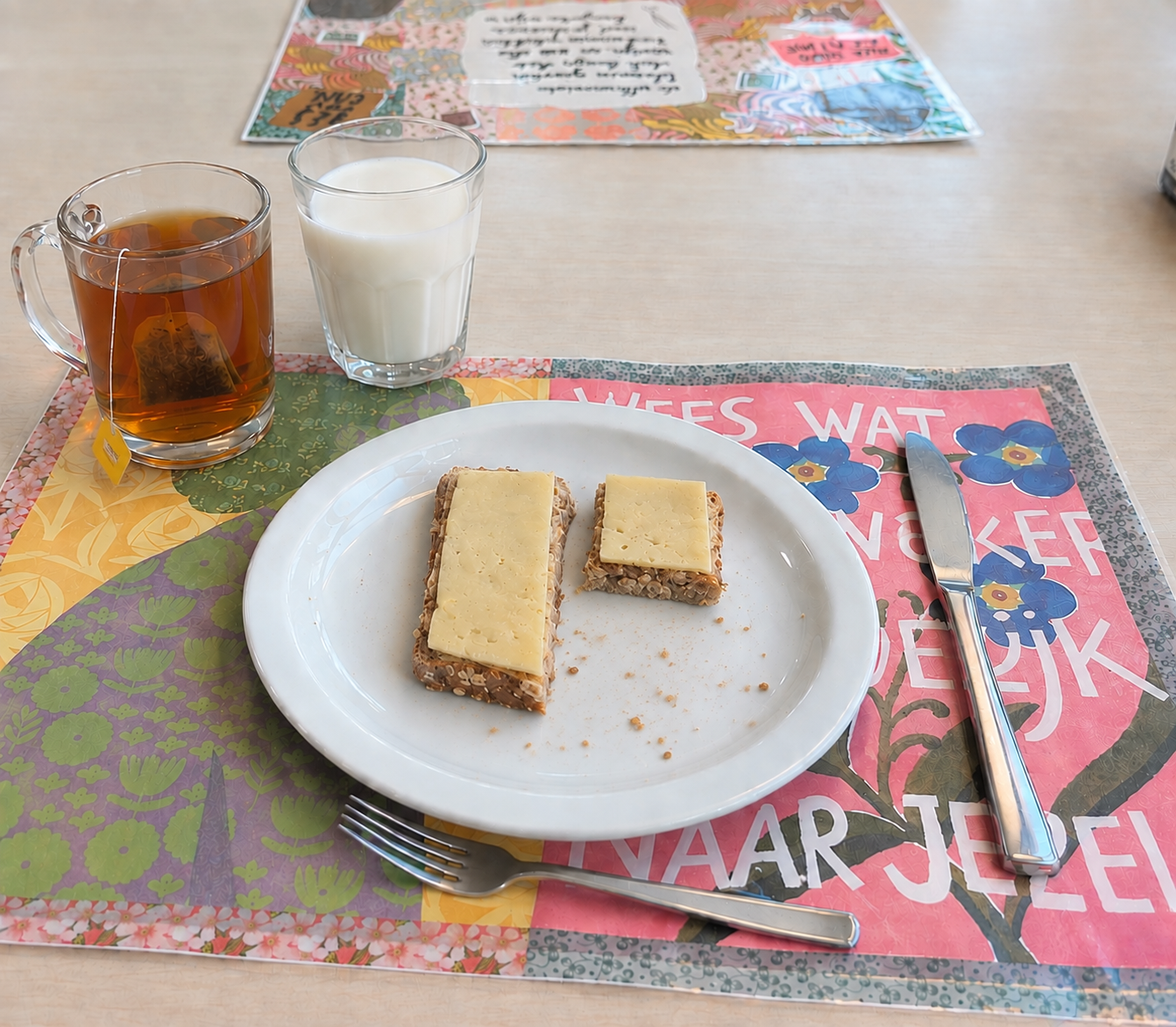 Twee boterhammen met kaas op bord, naast thee, melkglas, bestek en kleurrijke placemat. lunch, ontbijt, eetstoorniskliniek