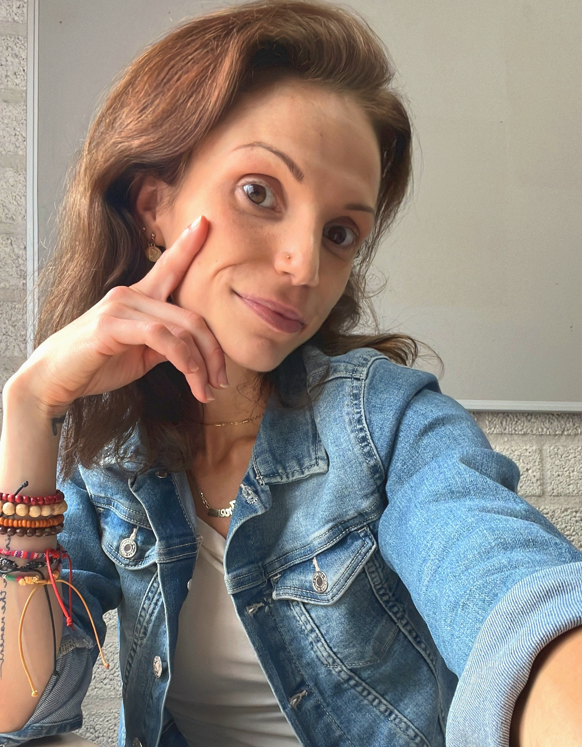 Portret van een vrouw met bruine haren, denim jas, hand bij gezicht, zittend tegen lichte muur.