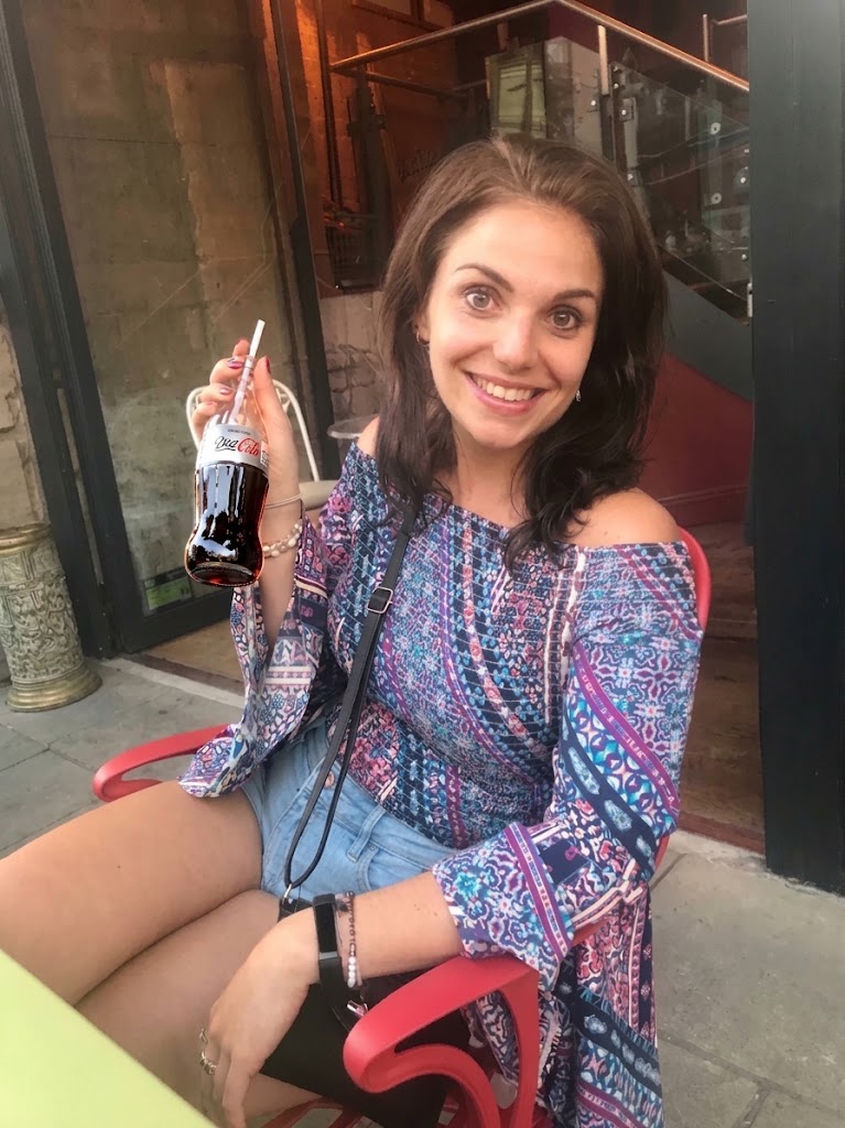 Glimlachende vrouw zit buiten en houdt een glas cola met rietje, kleurrijke jurk en tas. Uiteten, diner, restaurant met een eetstoornis