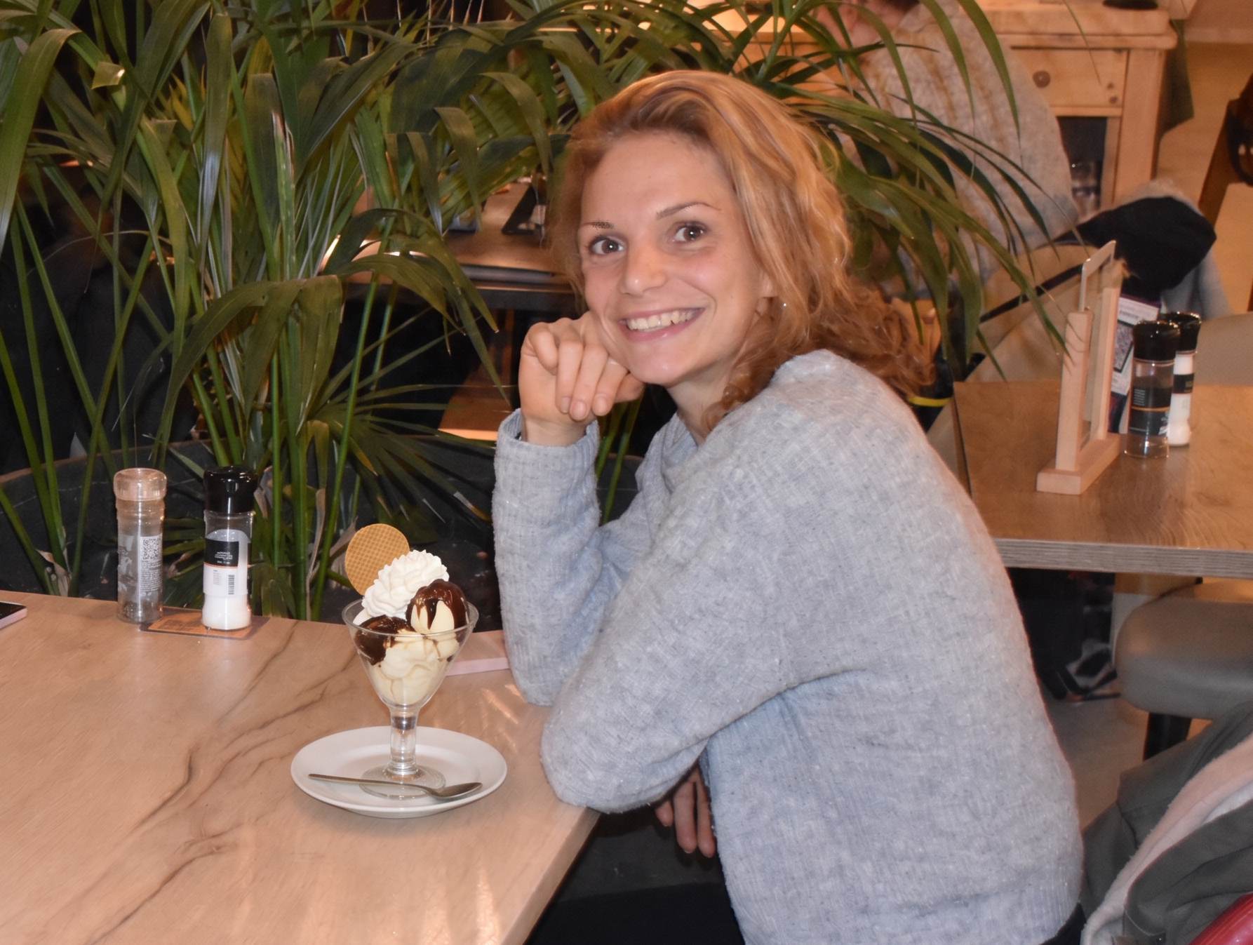 Glimlachende vrouw leunt aan houten bar met ijsdessert en kruidenpotjes, groene planten op achtergrond. Dame blanche. Uiteten, diner, restaurant met een eetstoornis