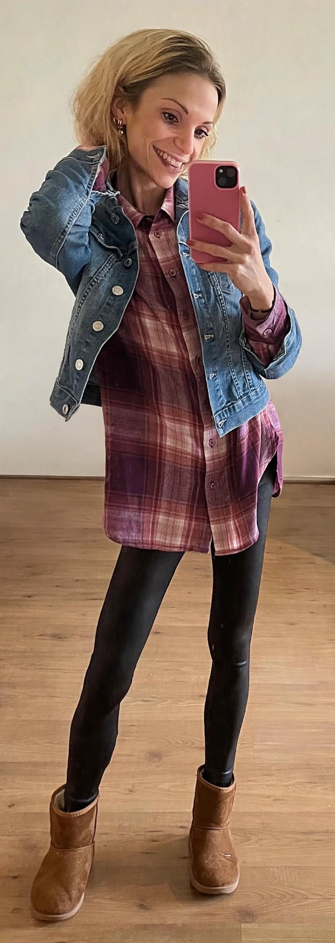Glimlachende vrouw in spiegelselfie met roze telefoon, blauwe jeansjas en ruitjesjurk in een woonkamer.