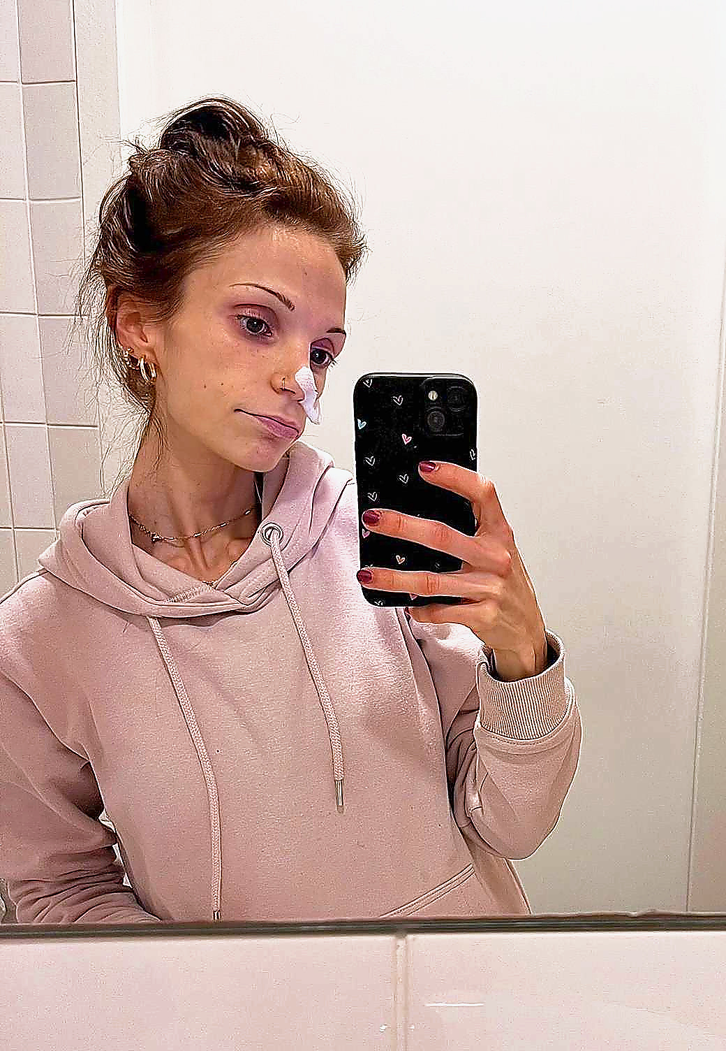 Jonge vrouw in beige hoodie maakt spiegel-selfie met smartphone, staand voor witte betegelde muur. Opname PAAZ ziekenhuis, sondevoeding, eetstoornis, anorexia, ondergewicht