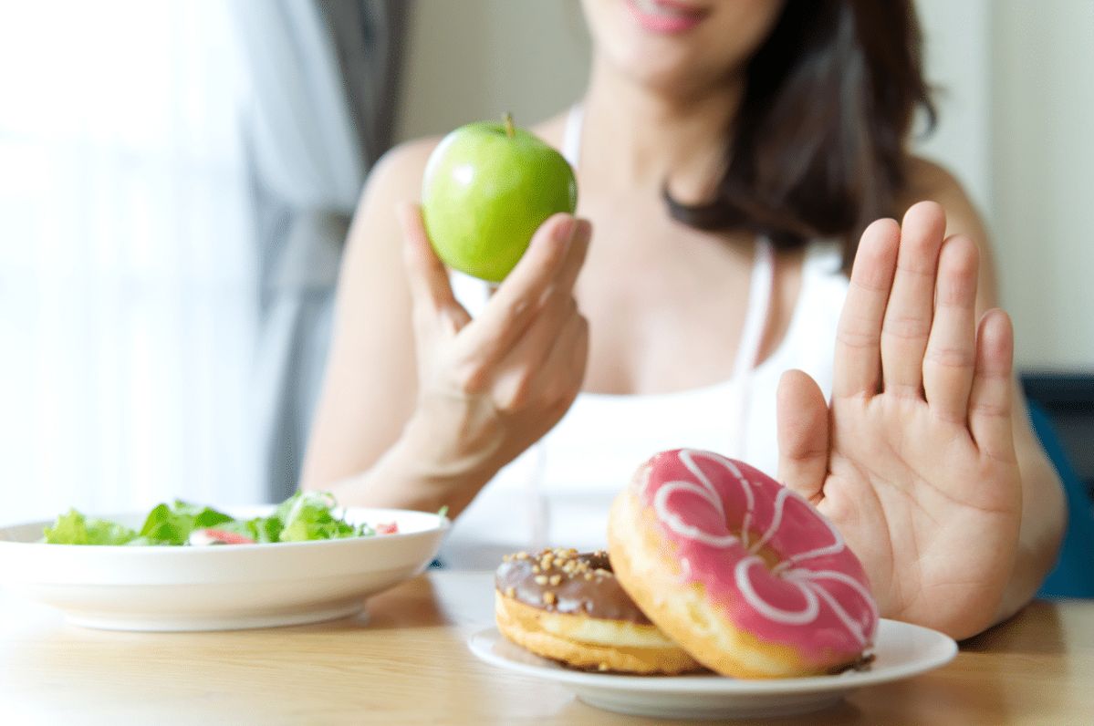 Vrouw houdt groene appel vast en toont handgebaar, met donut op tafel voor haar.