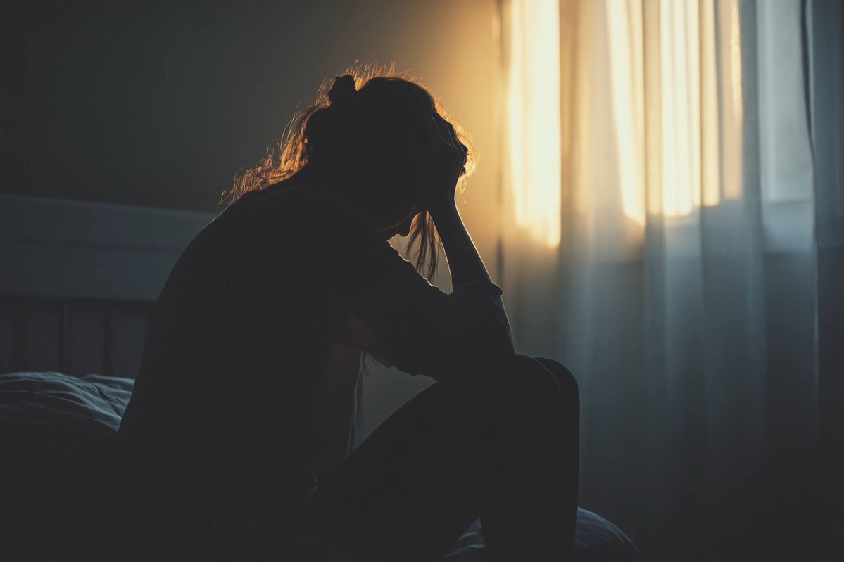 Zittende vrouw in silhouet, hoofd gebogen, bij raam met warm zonlicht en gordijnen.