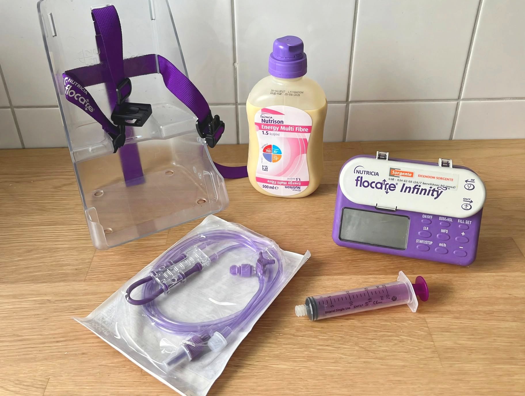 Paarse voedingsset met sondevoedingspomp, fles Nutricia en spuit op houten tafel, verpakkingen en slangen zichtbaar.