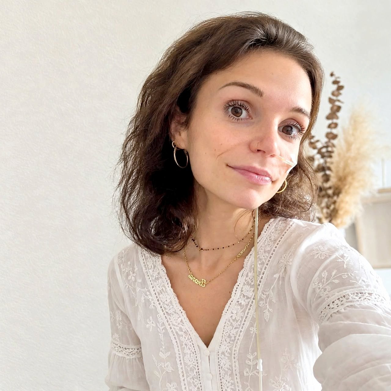 Portret van een vrouw met krullend bruin haar, witte blouse en gouden sieraden, glimlachend bij raam.