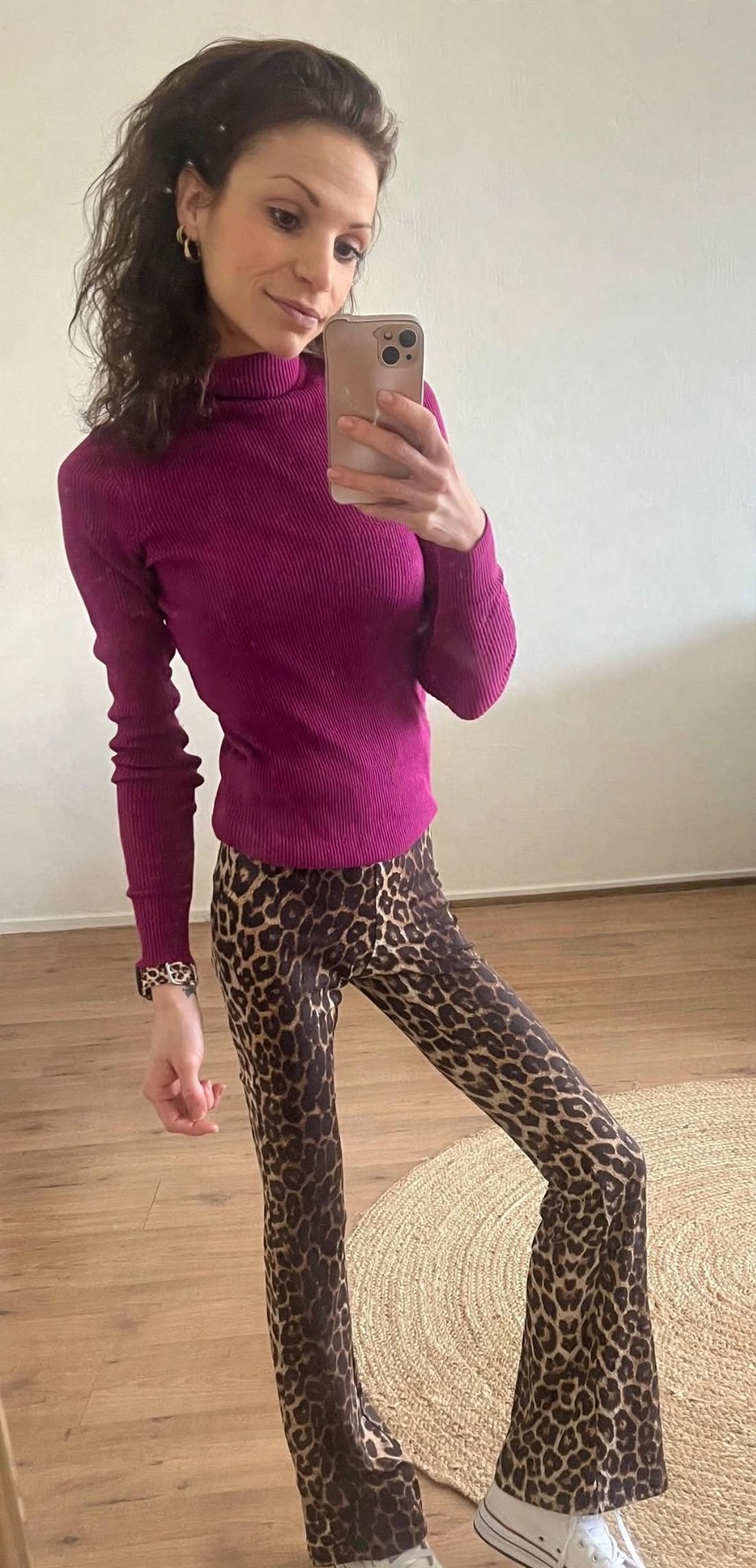Vrouw in spiegelselfie met paarse trui en luipaardprint broek, telefoon in hand, woonkamer op achtergrond.