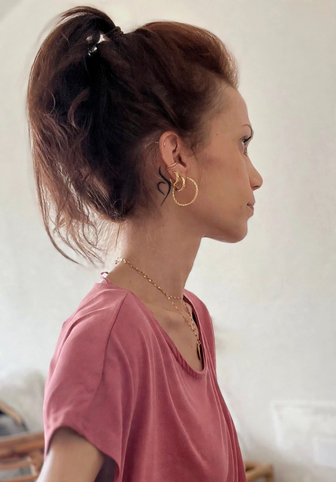 Zijaanzicht van vrouw met bruine knot, gouden oorbellen en ketting, rode top tegen witte achtergrond. Tattoage van het neda-symbol achter oor. National eating disorder association.