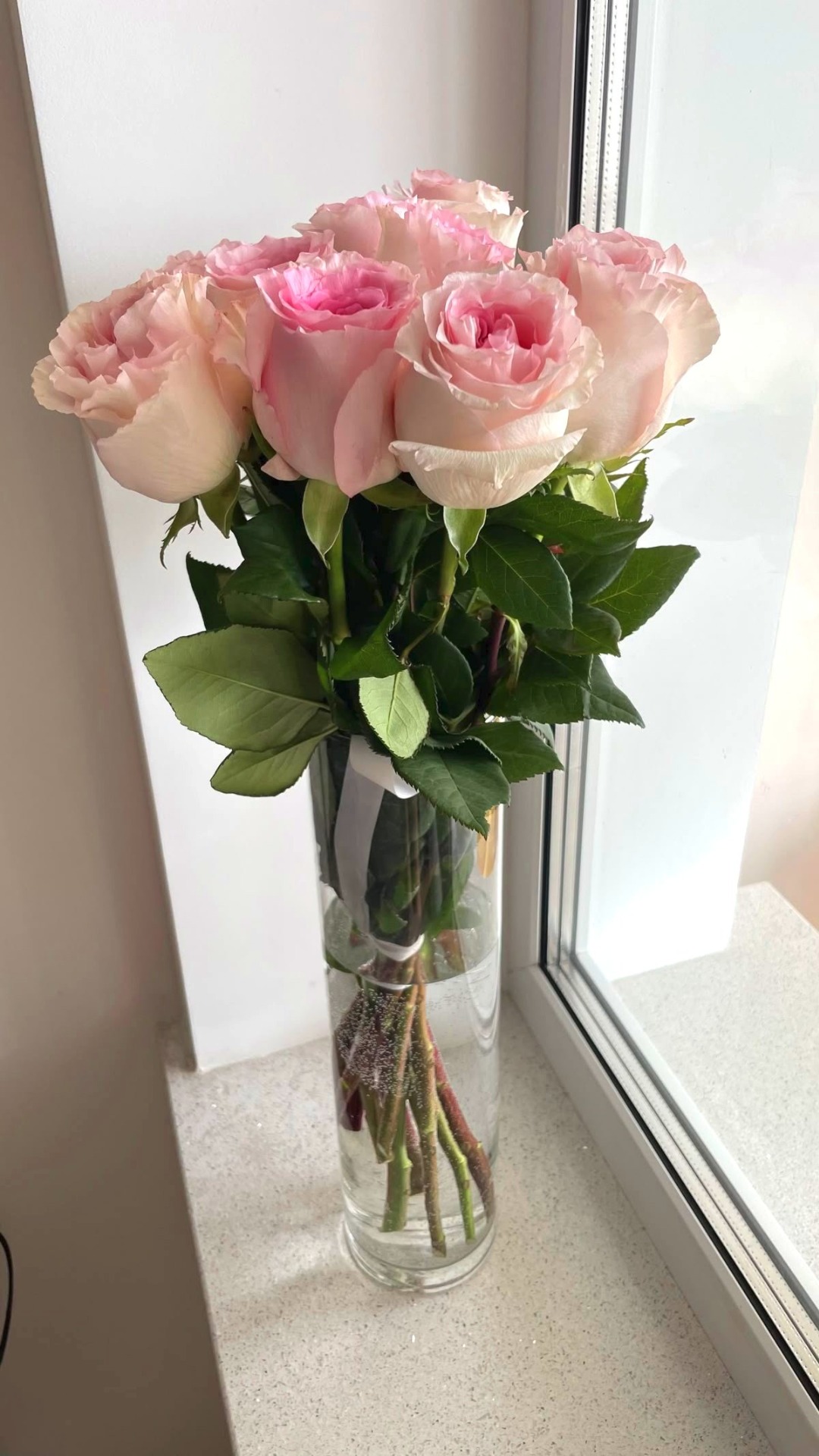 Roze en witte rozen in een glazen vaas met groene bladeren bij een raam.